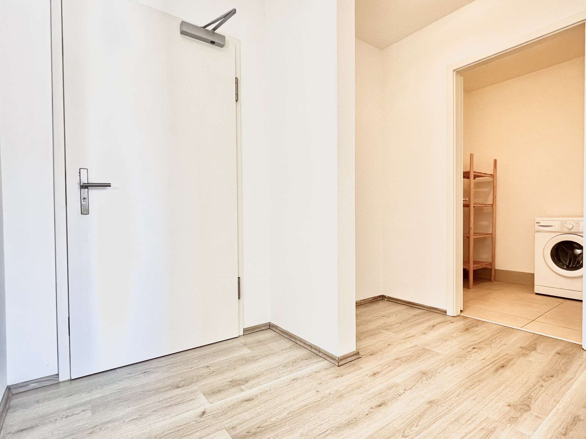 White hallway with closed white doors on the left and an open doorway to a laundry room on the right, showing a washing machine and a wooden shelf unit.