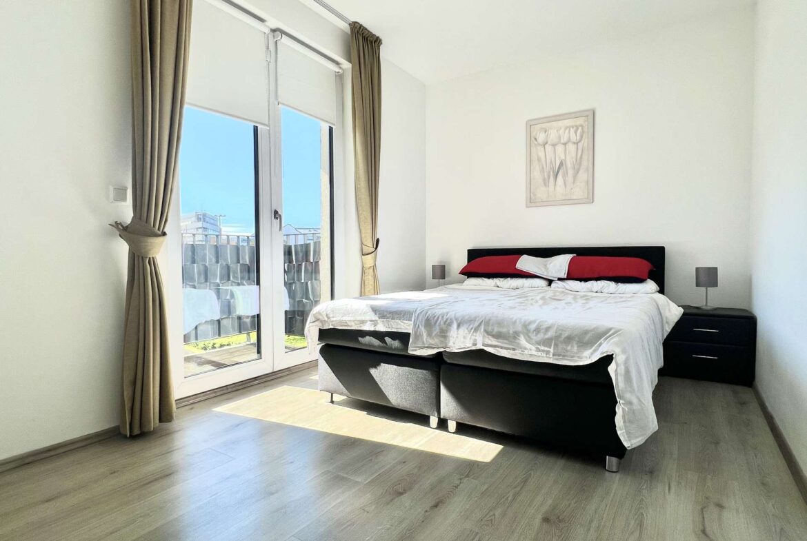 Bright, minimalist bedroom with a black platform bed, white bedding and red pillows, flanked by nightstands and lamps, next to glass balcony doors with beige curtains.