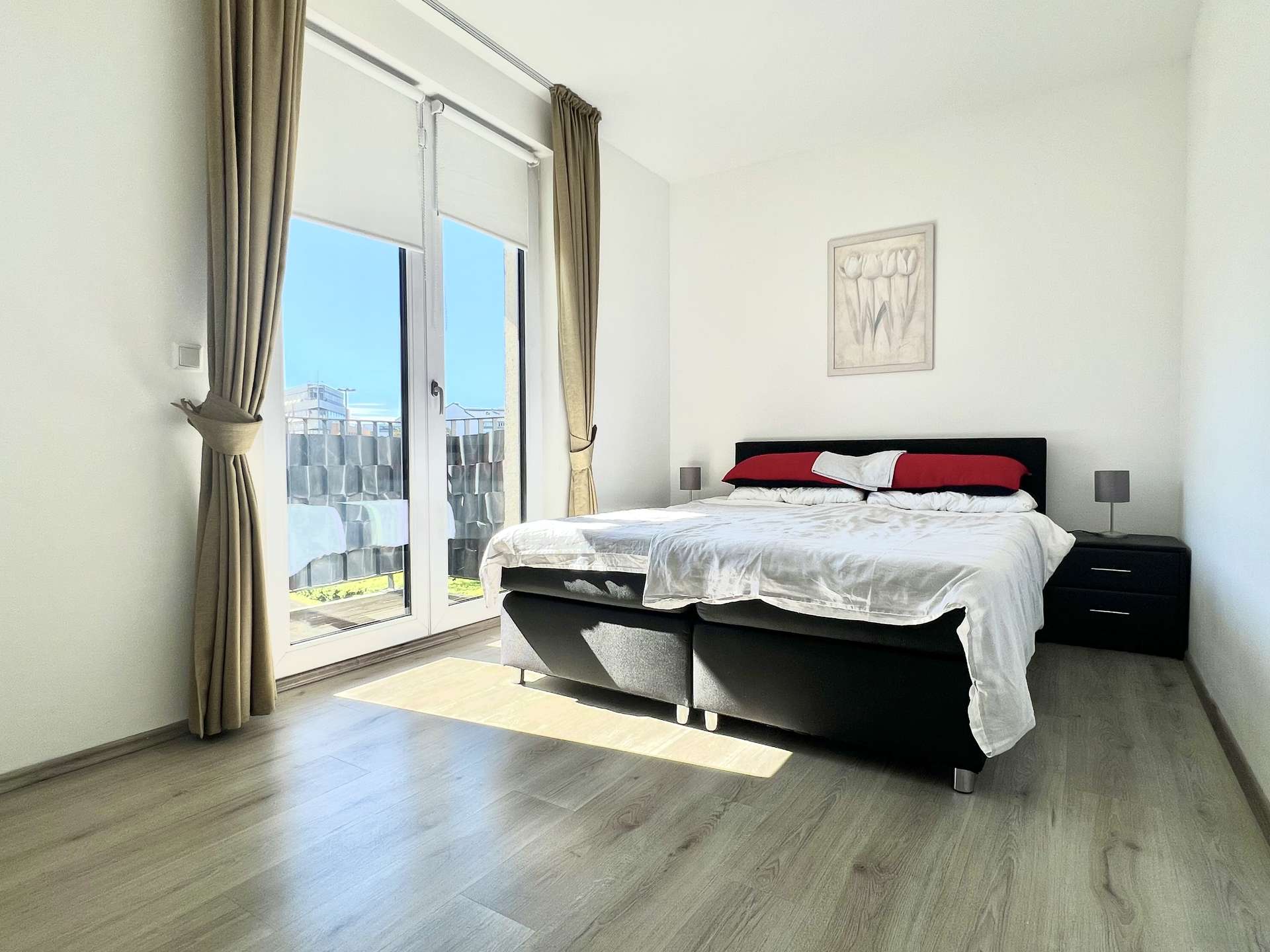 Bright, minimalist bedroom with a black platform bed, white bedding and red pillows, flanked by nightstands and lamps, next to glass balcony doors with beige curtains.