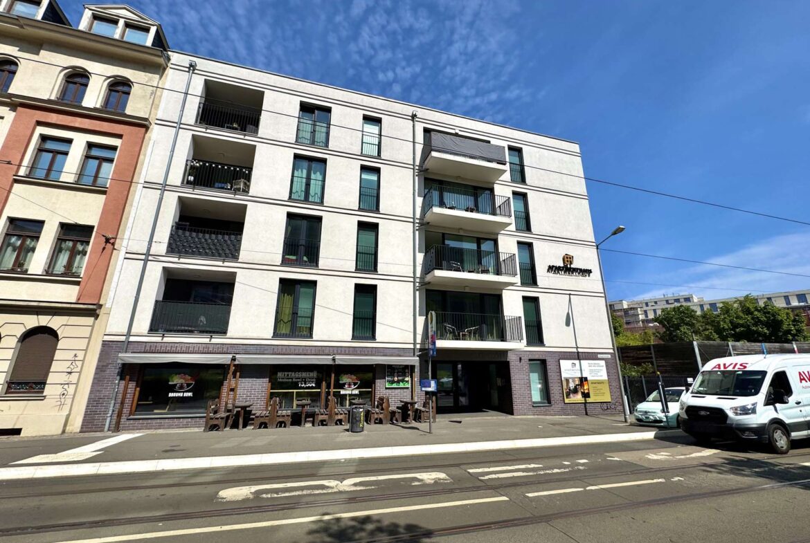 Modern white apartment building with balconies on a sunny street; tram tracks run along the foreground and a few vehicles nearby.