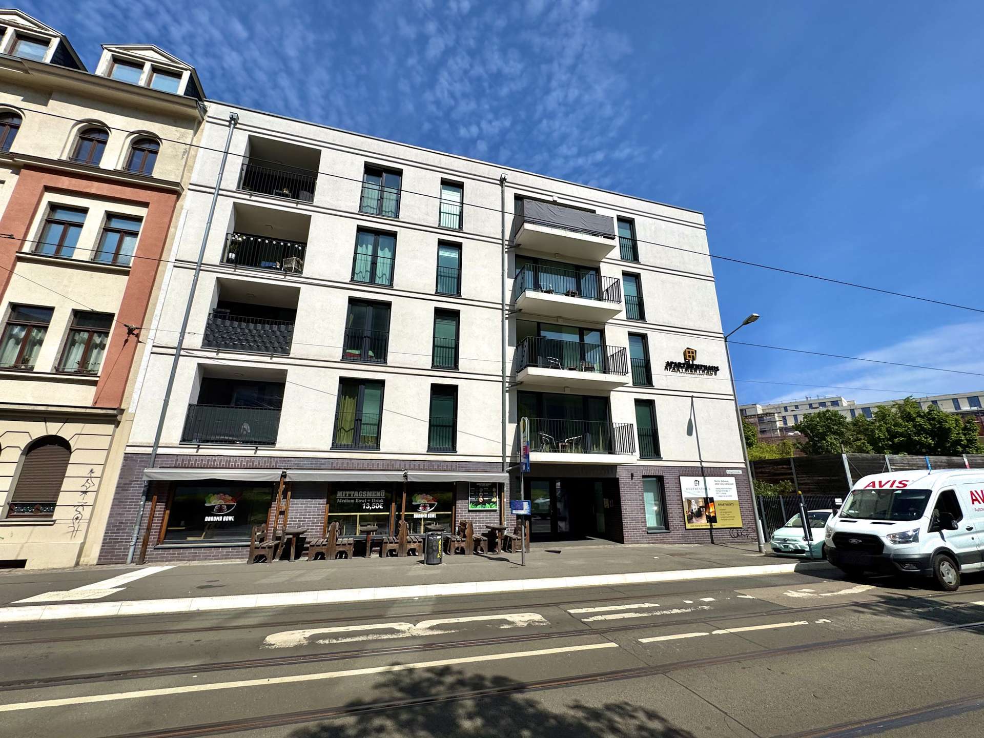 Modern white apartment building with balconies on a sunny street; tram tracks run along the foreground and a few vehicles nearby.