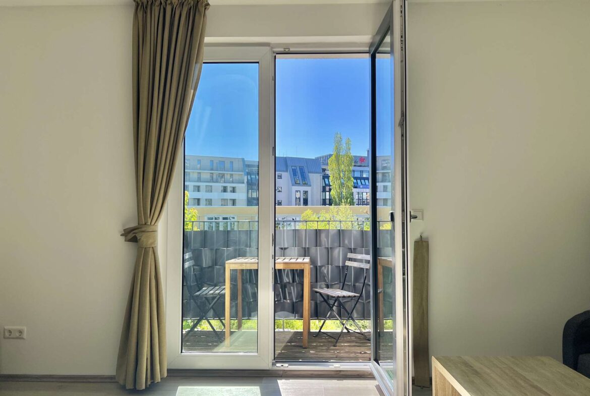 Open sliding glass balcony door in a bright room with beige curtains; outside there is a small table and chairs on a deck, urban buildings and a blue sky visible.