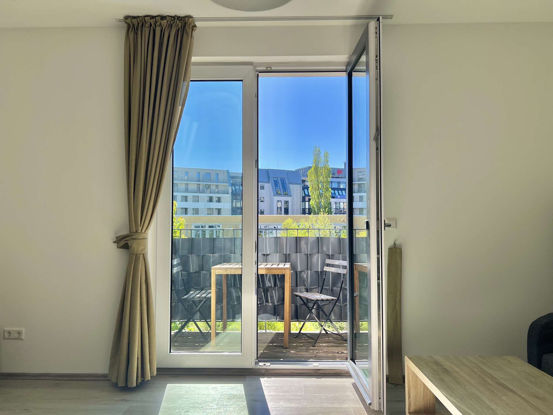 Open sliding glass balcony door in a bright room with beige curtains; outside there is a small table and chairs on a deck, urban buildings and a blue sky visible.