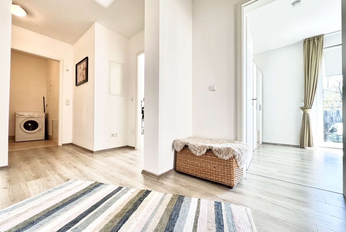 Bright open living space with a striped rug, wicker bench with fur throw, and a laundry area with a washing machine on the left.