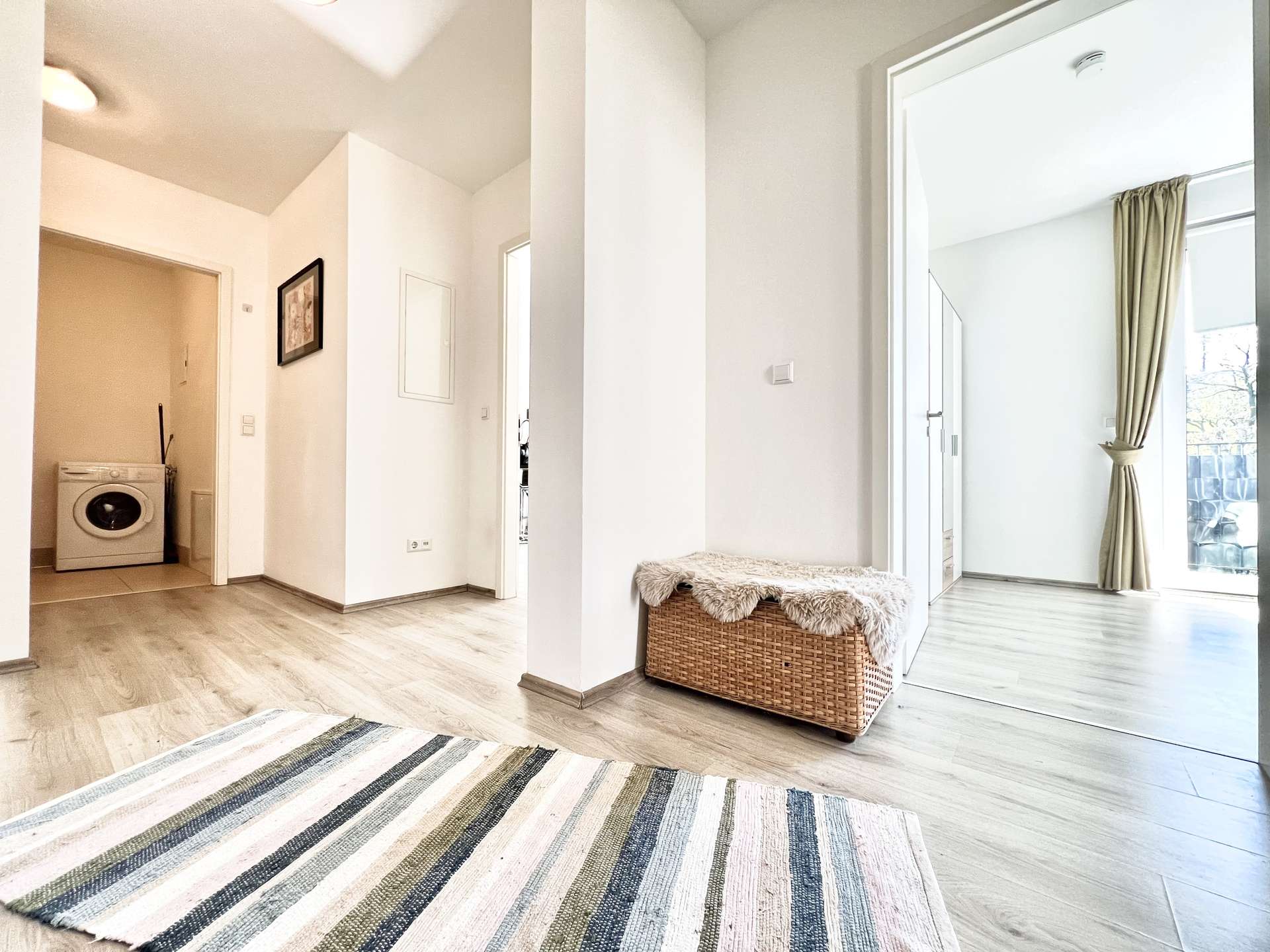 Bright open living space with a striped rug, wicker bench with fur throw, and a laundry area with a washing machine on the left.