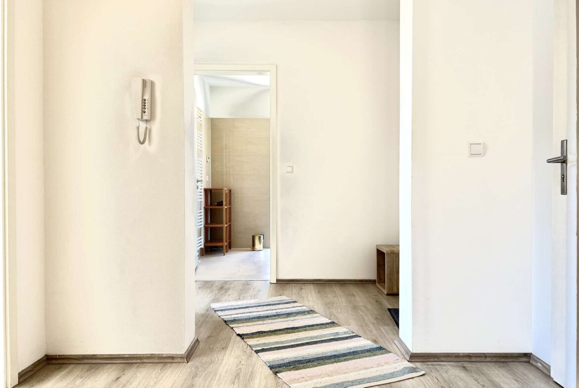 Bright hallway with light wood floor, striped rug, and doorway into a tiled room with a small wooden shelf visible.