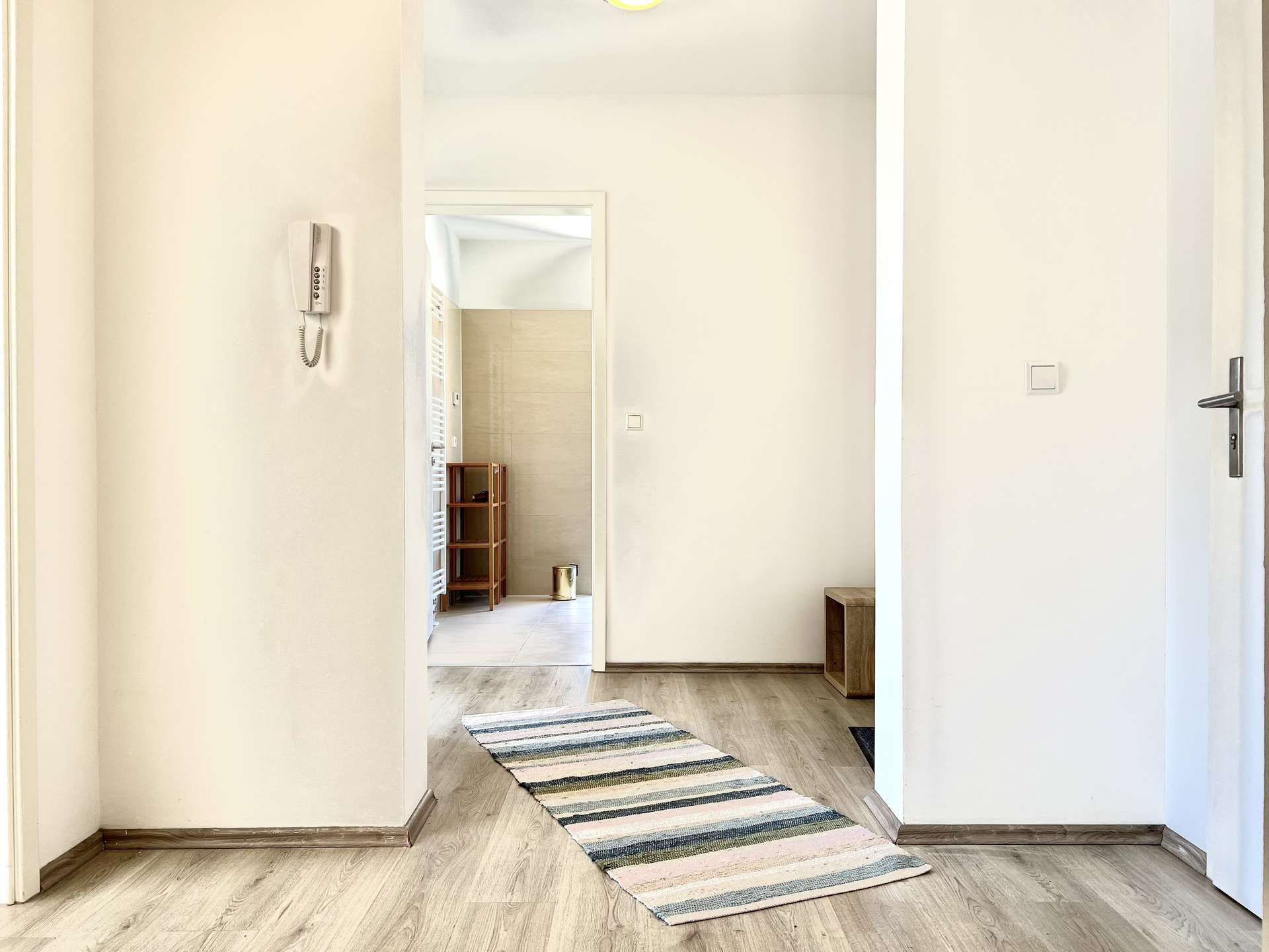 Bright hallway with light wood floor, striped rug, and doorway into a tiled room with a small wooden shelf visible.
