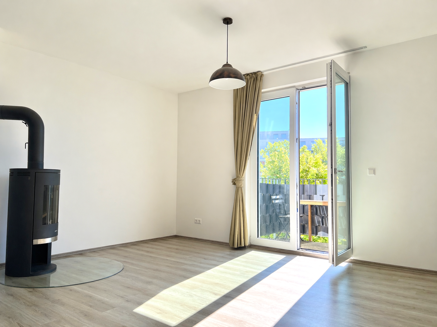 Bright, minimalist living room with a black modern stove on the left, and an open glass door to a sunny balcony outside.
