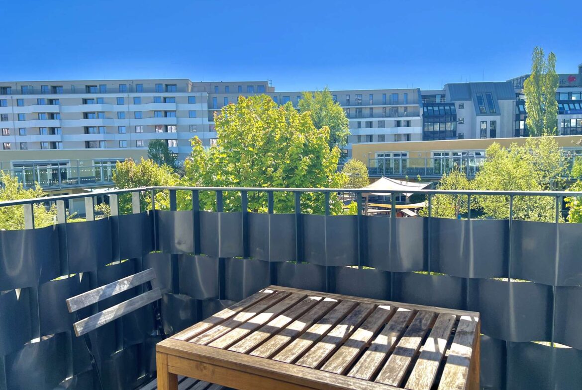 Wooden outdoor table and bench on a balcony with a dark metal railing, overlooking modern buildings and green trees under a bright blue sky.