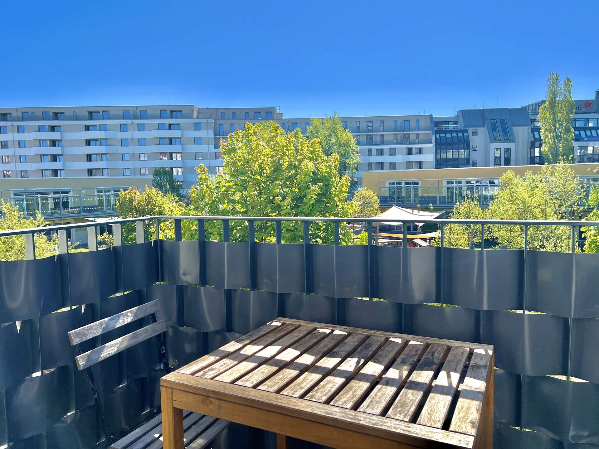 Wooden outdoor table and bench on a balcony with a dark metal railing, overlooking modern buildings and green trees under a bright blue sky.