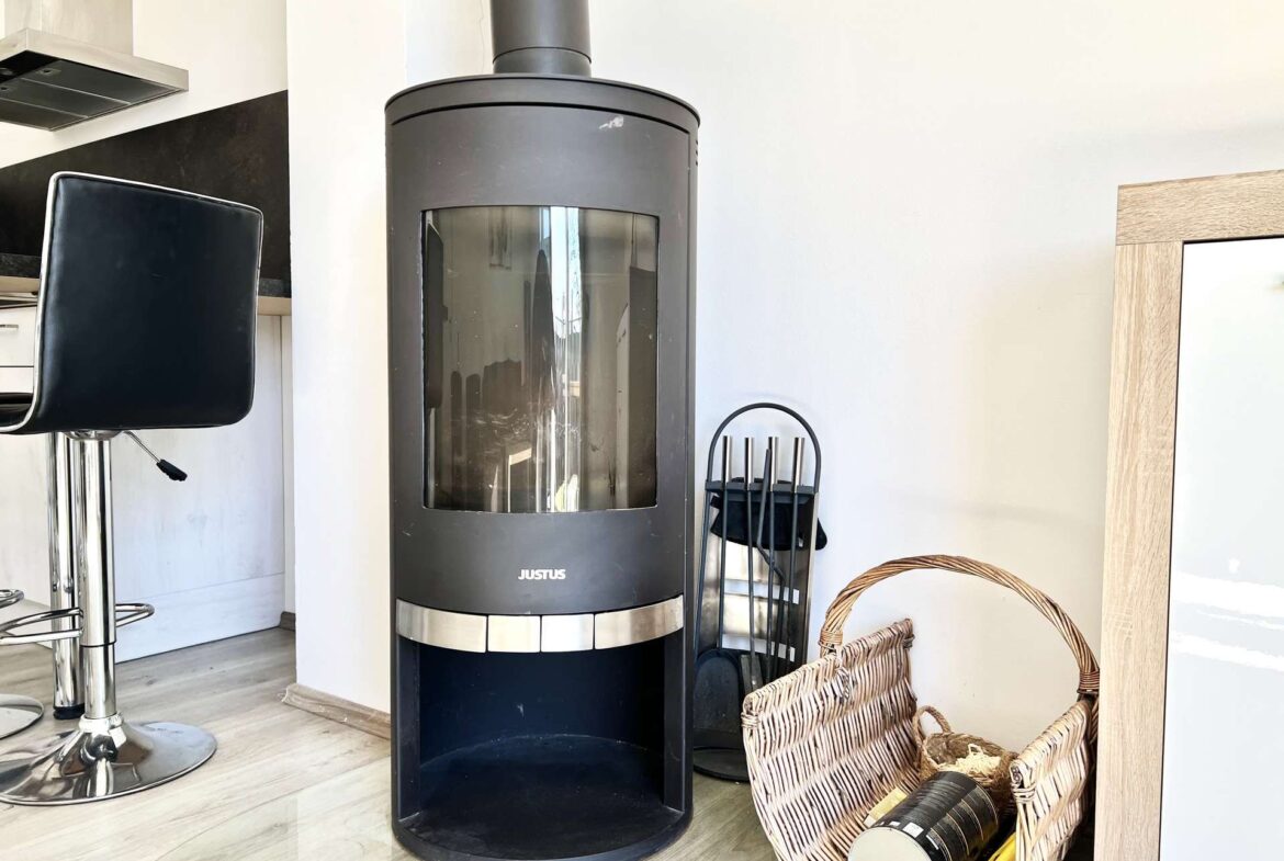 Tall black cylindrical wood stove with glass window in a bright living area, with baskets and fireplace tools nearby