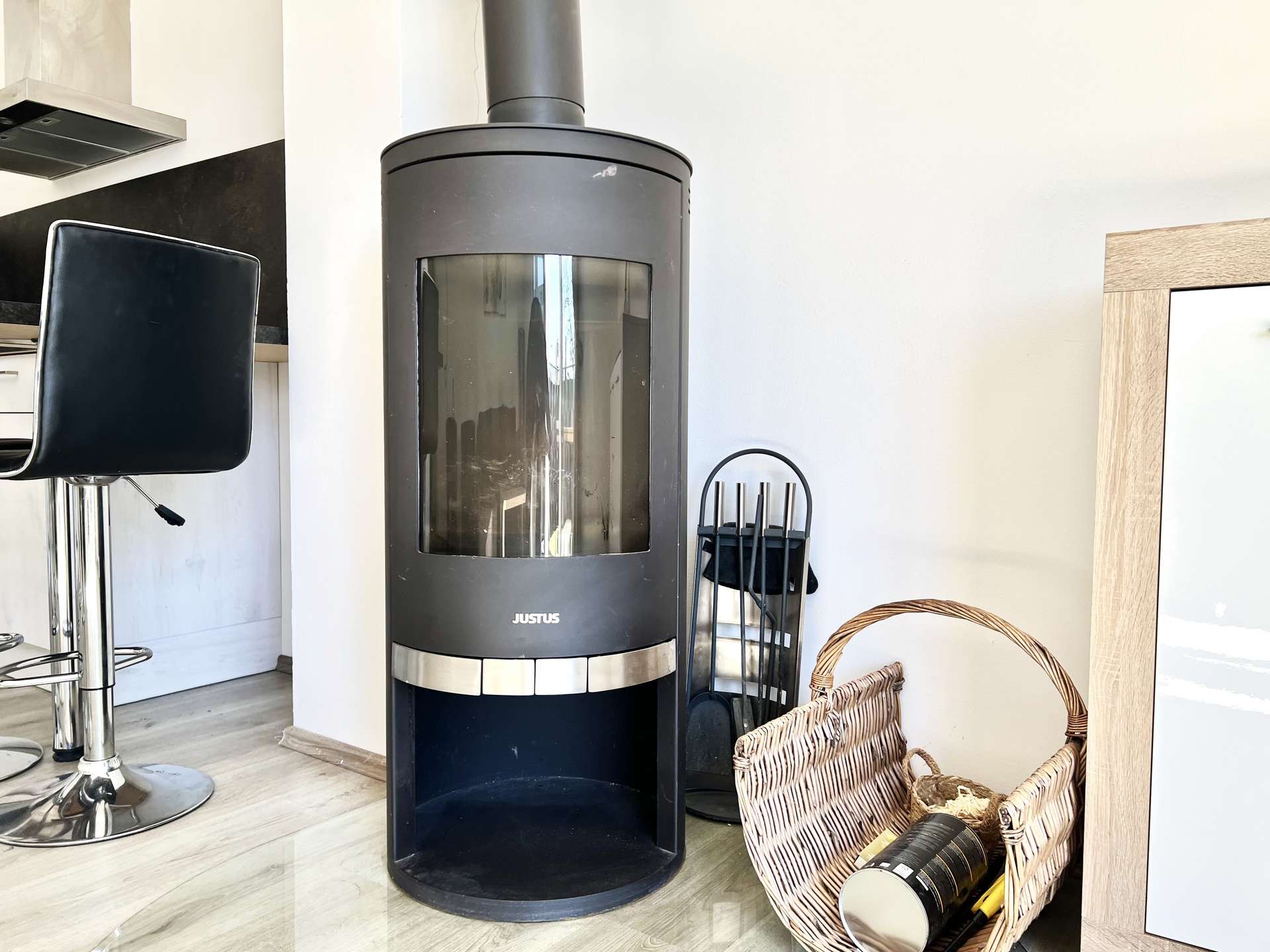 Tall black cylindrical wood stove with glass window in a bright living area, with baskets and fireplace tools nearby