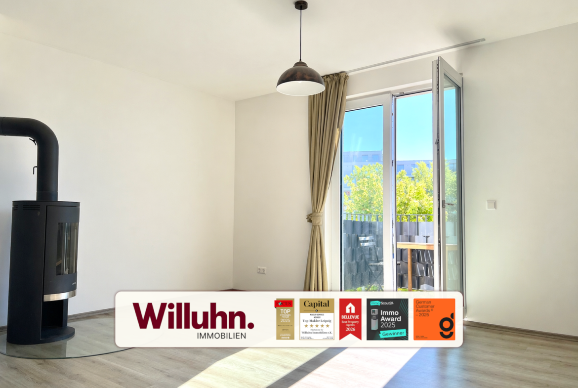Bright living room with a black wood stove on the left, large glass balcony door, beige curtains, and a Willuhn Immobilien banner across the bottom.