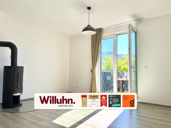 Bright living room with a black wood stove on the left, large glass balcony door, beige curtains, and a Willuhn Immobilien banner across the bottom.