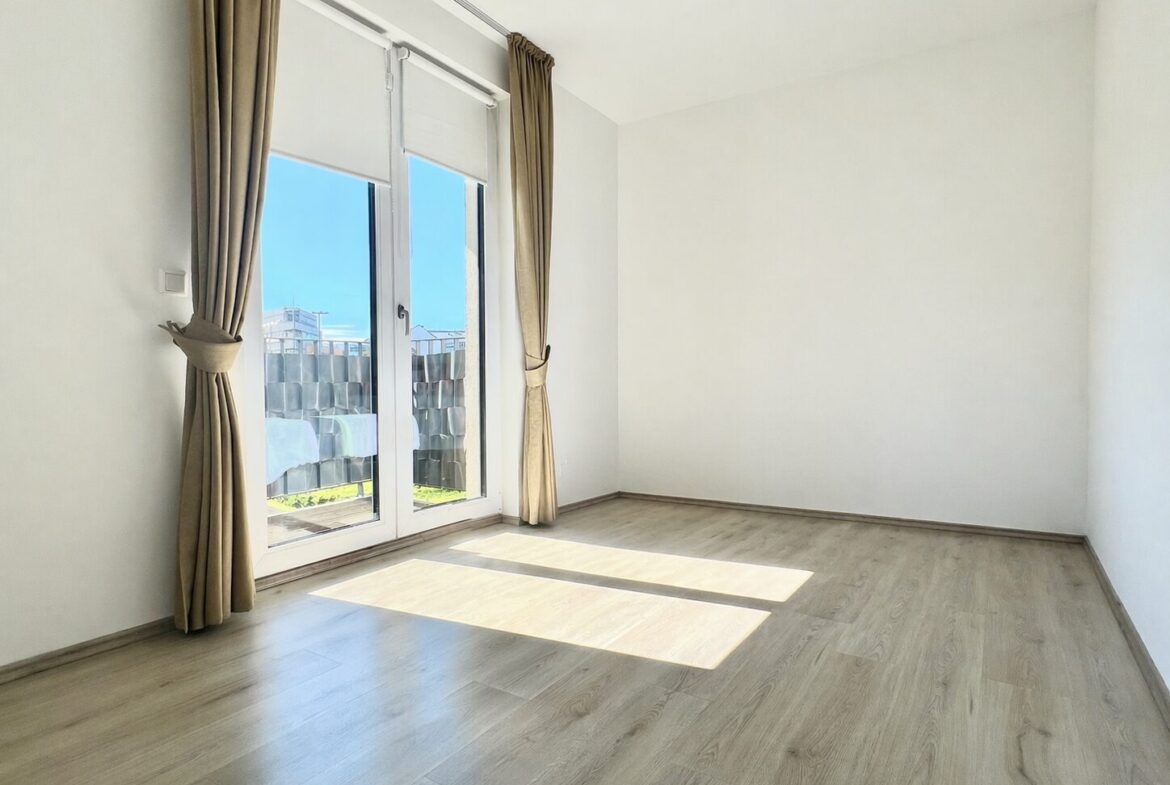 Bright empty room with large glass doors and beige curtains, wooden floor, sunlight streaming in.