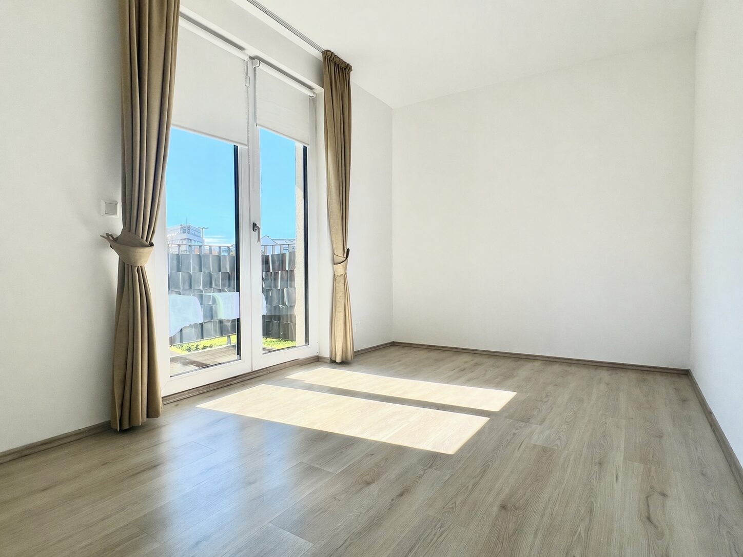 Bright empty room with large glass doors and beige curtains, wooden floor, sunlight streaming in.