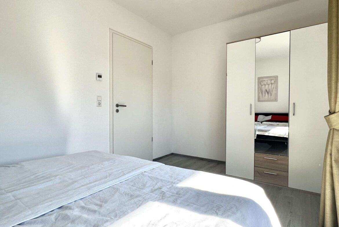 A bright hotel-style bedroom with white walls, a large mirrored wardrobe, and a bed in the foreground.