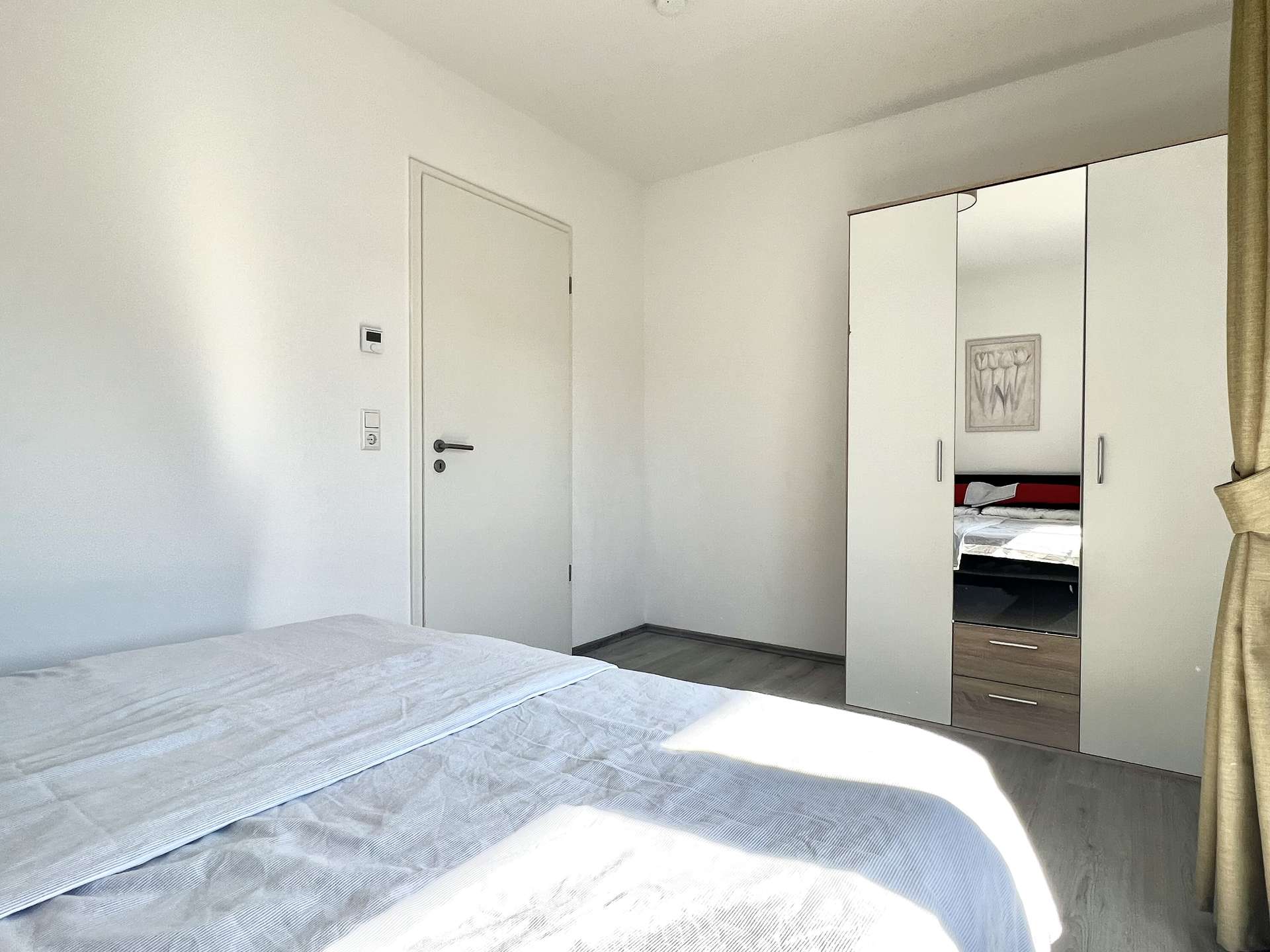 A bright hotel-style bedroom with white walls, a large mirrored wardrobe, and a bed in the foreground.
