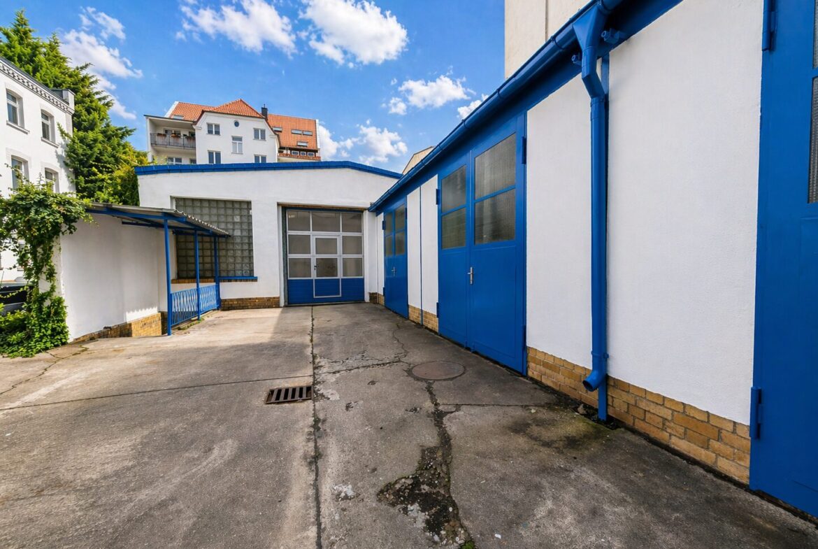 White industrial building with blue doors and trim in a paved courtyard under a blue sky to the left and a garage door in the center-left.