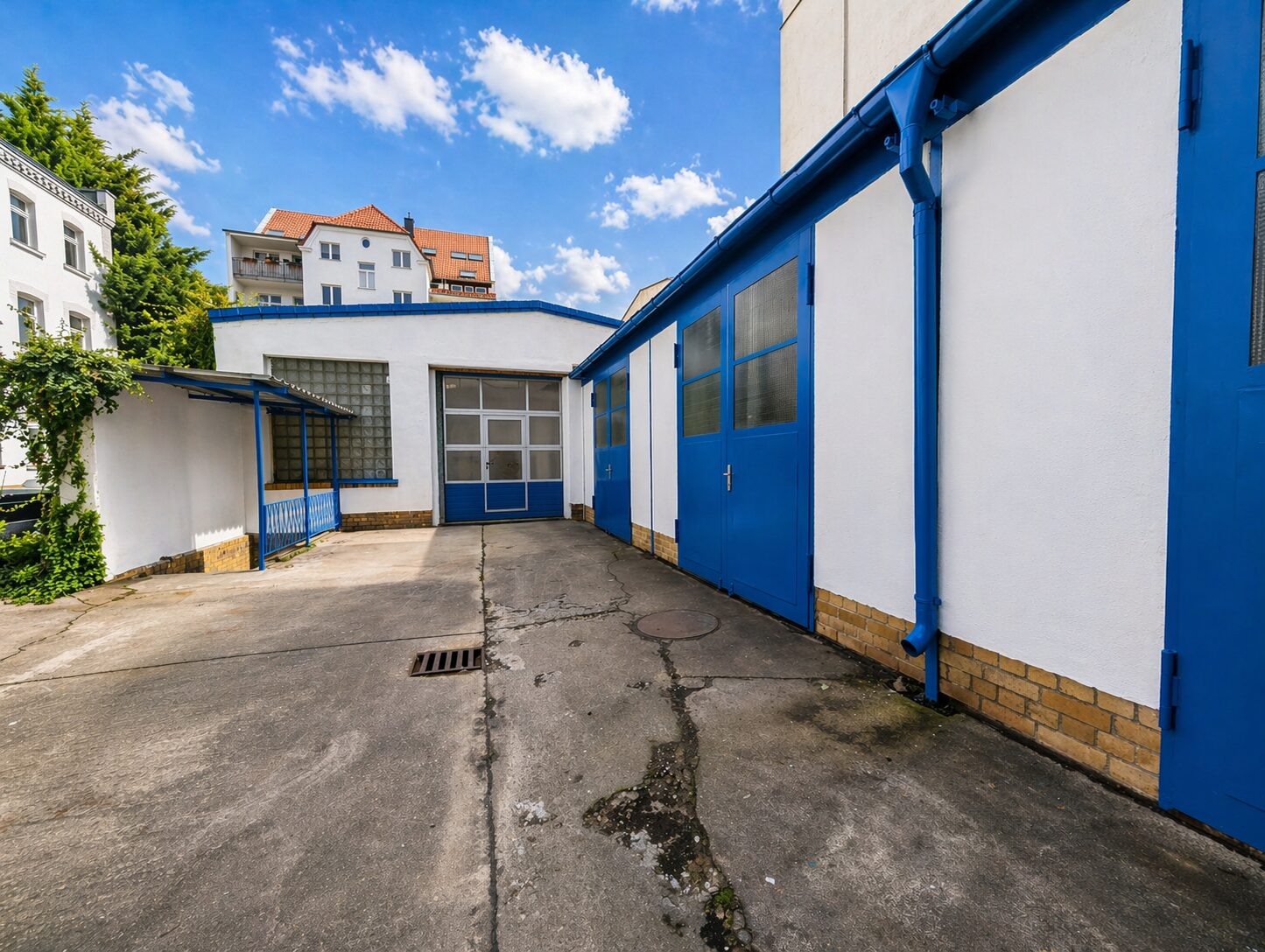 White industrial building with blue doors and trim in a paved courtyard under a blue sky to the left and a garage door in the center-left.