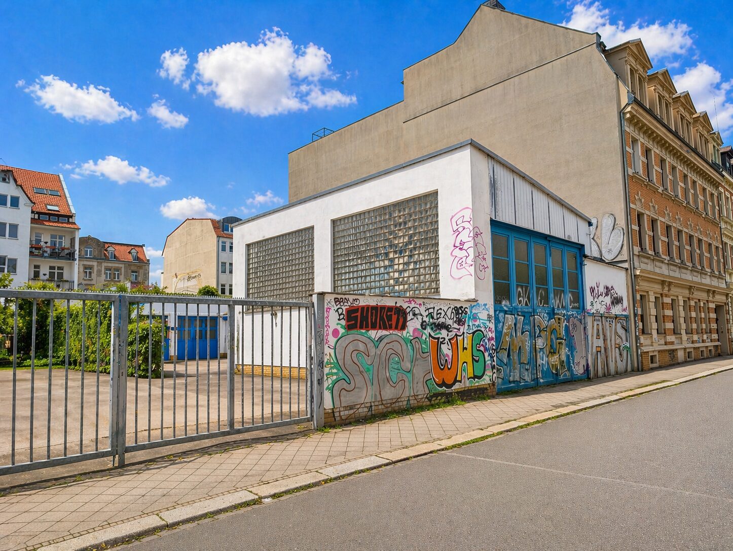 Urban street scene with a graffiti-covered wall, metal fence gate, and nearby beige/white buildings under a blue sky.