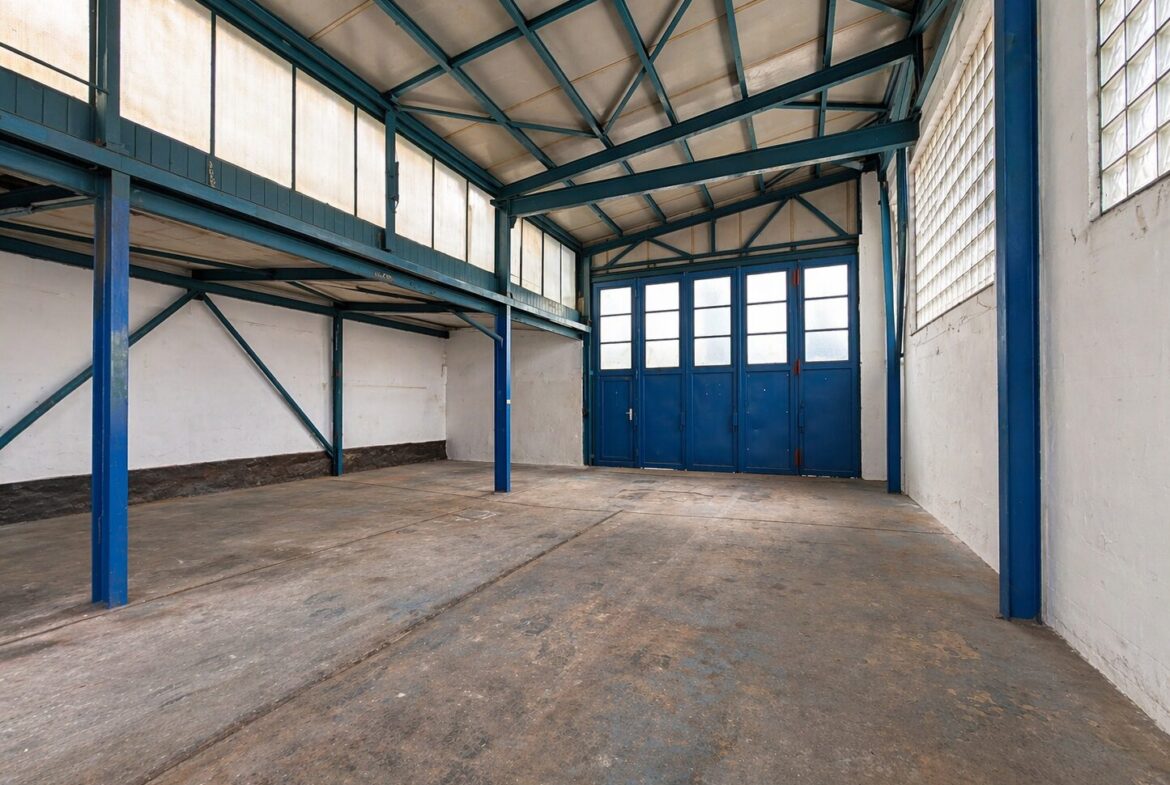Empty industrial warehouse interior with blue steel beams and a large blue door at the far end. The concrete floor is worn and the high ceiling has exposed trusses.