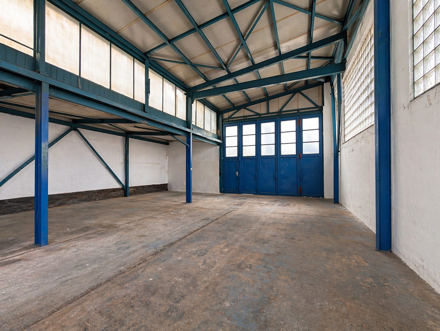Empty industrial warehouse interior with blue steel beams and a large blue door at the far end. The concrete floor is worn and the high ceiling has exposed trusses.