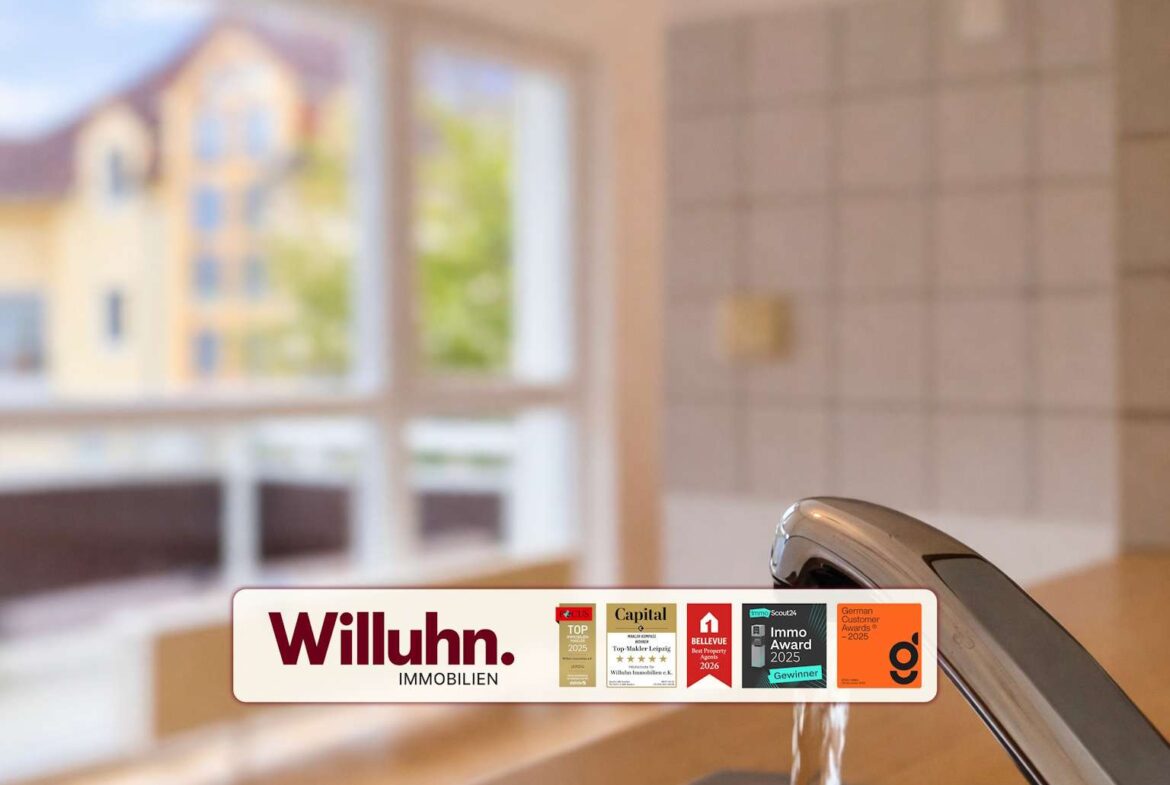 Close-up of a kitchen sink faucet with a Willuhn Immobilien promotional banner and awards displayed on the counter in the foreground.