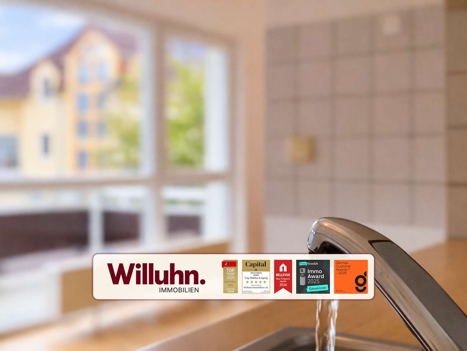 Close-up of a kitchen sink faucet with a Willuhn Immobilien promotional banner and awards displayed on the counter in the foreground.