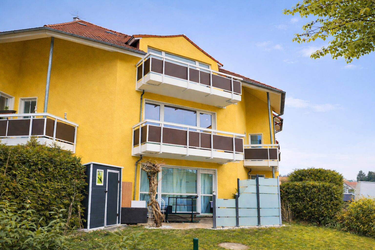 Yellow apartment building with white-framed brown balconies under a clear blue sky, and a small patio area in the foreground.
