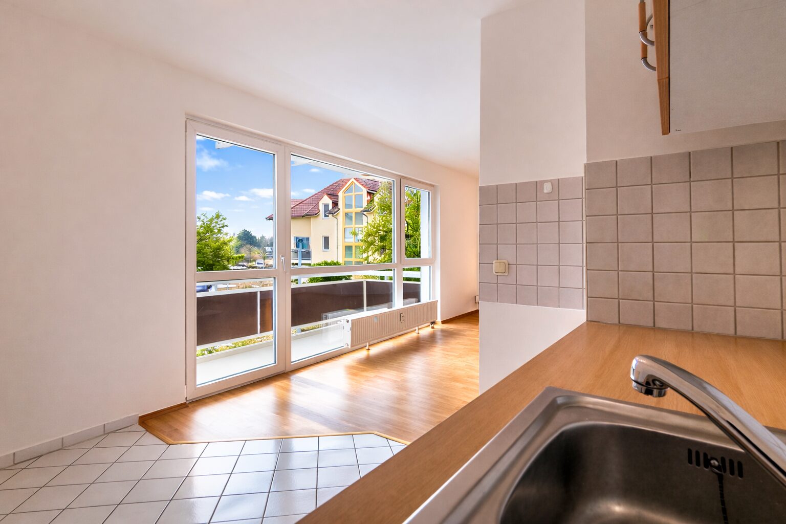 Bright kitchen with a stainless sink and wooden counter, tiled backsplash, and large glass doors opening to a sunny balcony with a street view.