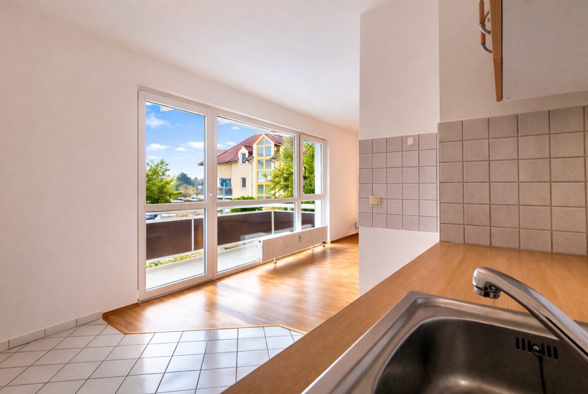 Bright kitchen with a stainless steel sink and wooden counter, large glass doors opening to a balcony with a sunny residential street outside.
