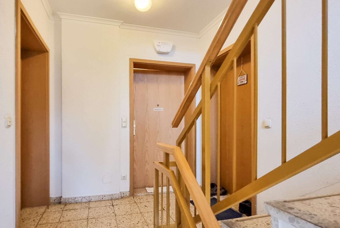 Interior stairwell with a wooden handrail on the right, tiled floor, white walls, and a doorway at the end.
