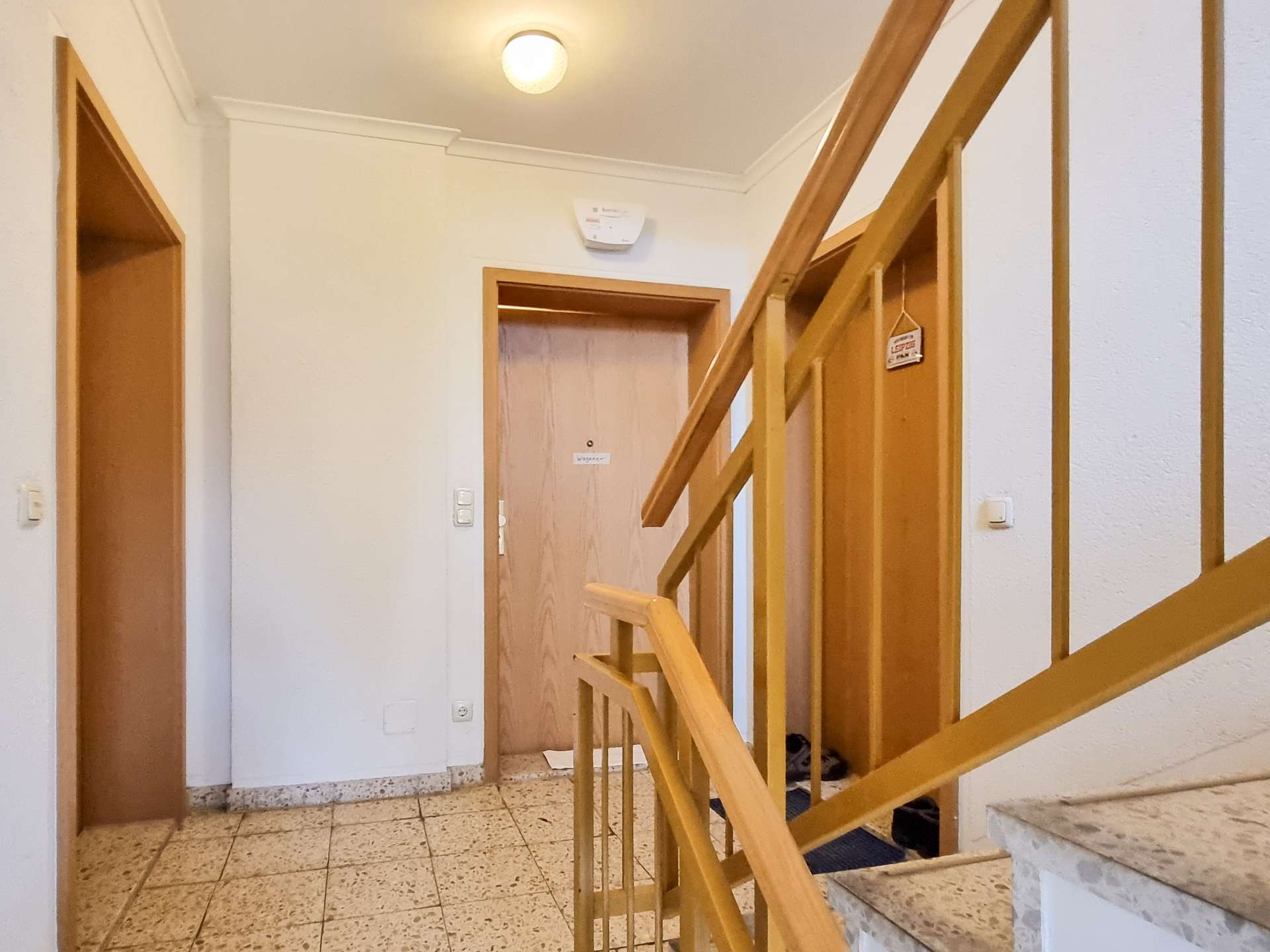 Interior stairwell with a wooden handrail on the right, tiled floor, white walls, and a doorway at the end.