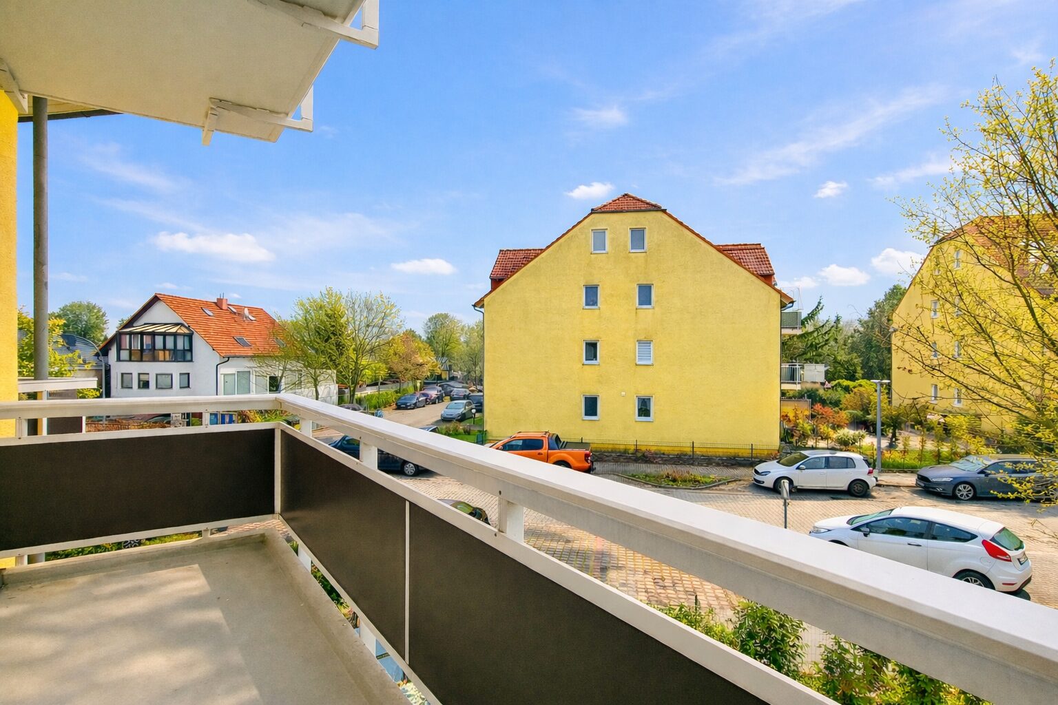 Balcony view of a sunny residential street with a large yellow apartment building across, several parked cars, and blue sky.