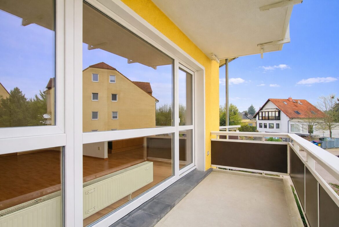 Balcony with glass doors opening to a yellow building; view of neighboring houses and blue sky.