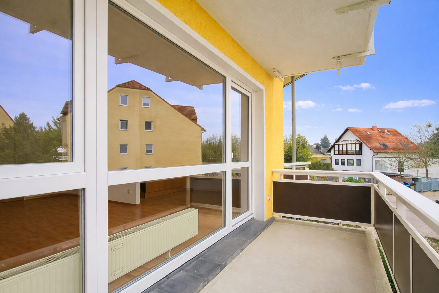 Balcony with glass doors opening to a yellow building; view of neighboring houses and blue sky.