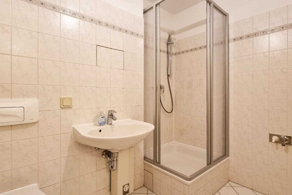 Small bathroom with a corner shower stall, white tiled walls, and a white wall-mounted sink with a faucet and soap bottle nearby