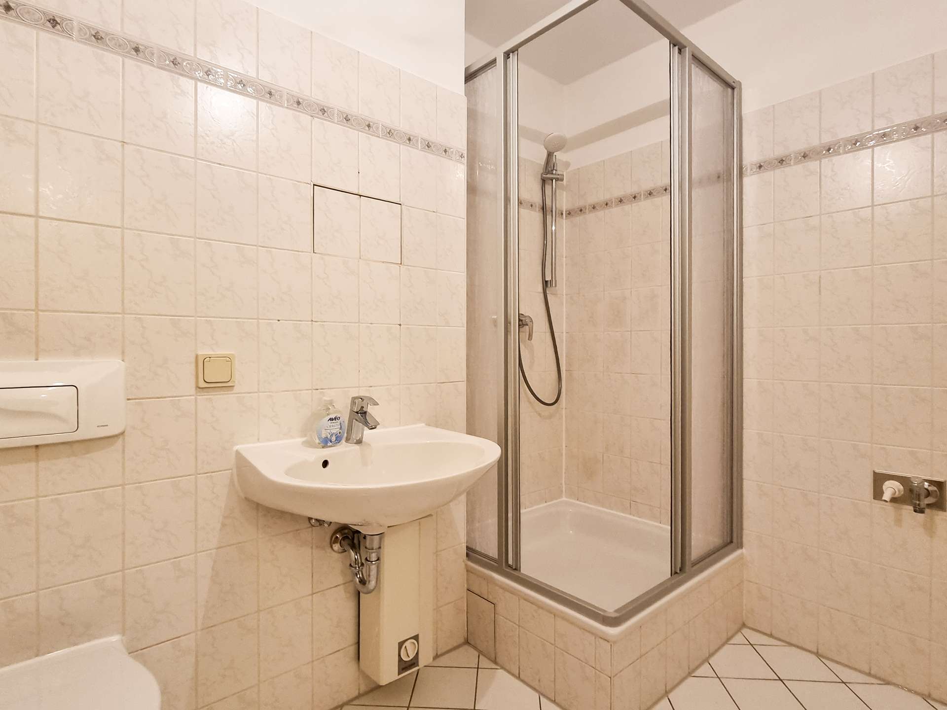 Small bathroom with a corner shower stall, white tiled walls, and a white wall-mounted sink with a faucet and soap bottle nearby