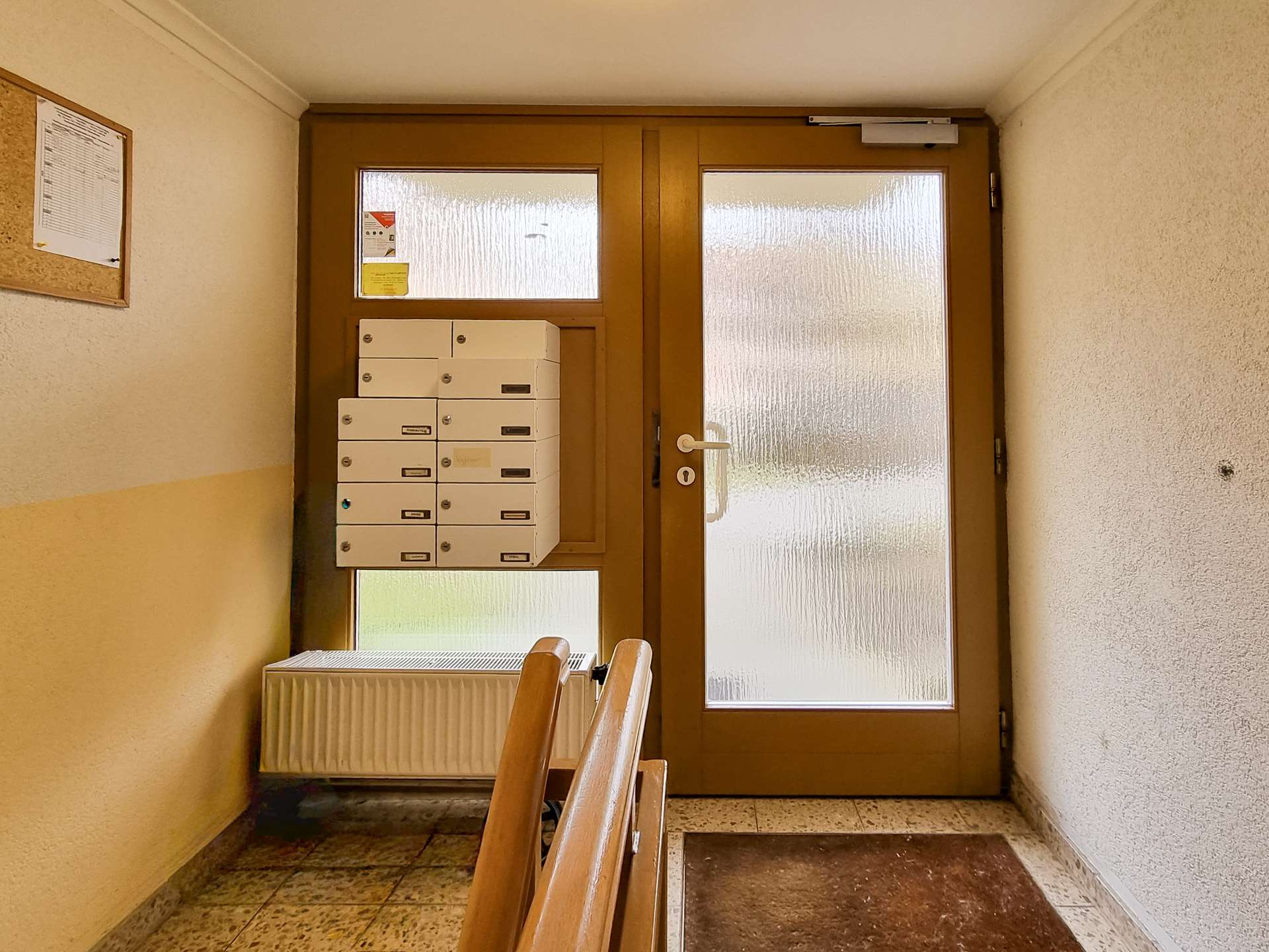 Interior stairwell with white mailboxes on the left, a brown wooden door with frosted glass panels, and a radiator below.