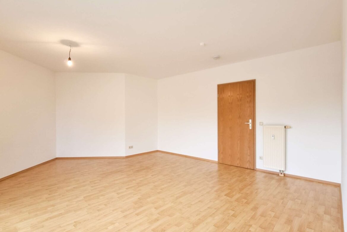 Empty, unfurnished room with a single hanging light bulb, wooden door on the right, and a radiator against the wall.