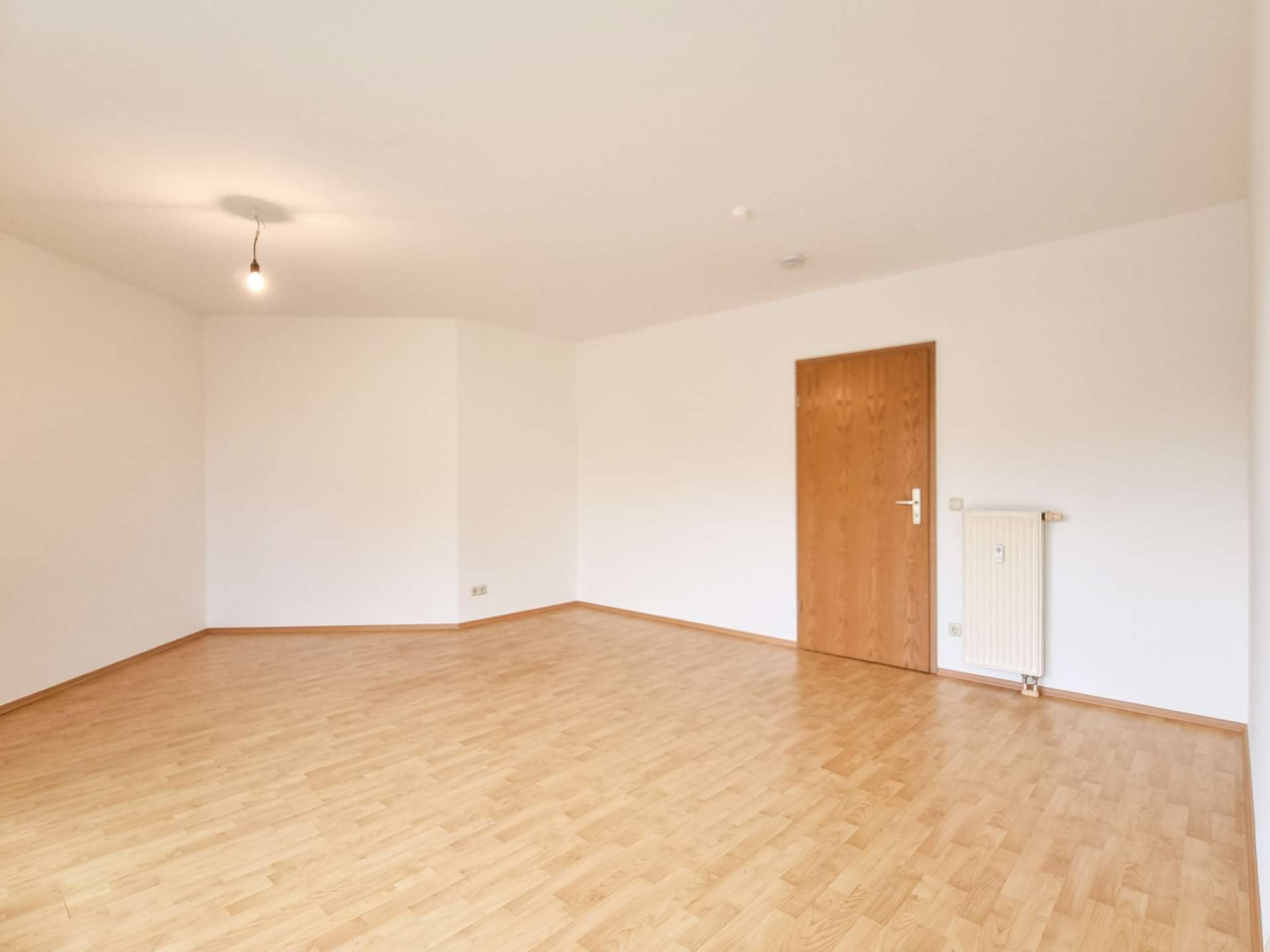 Empty, unfurnished room with a single hanging light bulb, wooden door on the right, and a radiator against the wall.