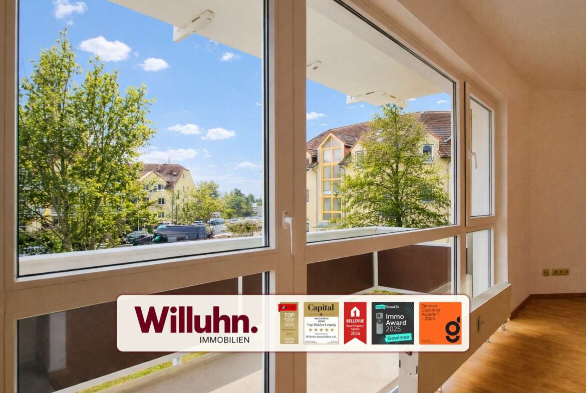 Bright living space with large glass windows showing a tree-filled street and yellow buildings outside; Willuhn Immobilien branding banner with awards along the bottom
