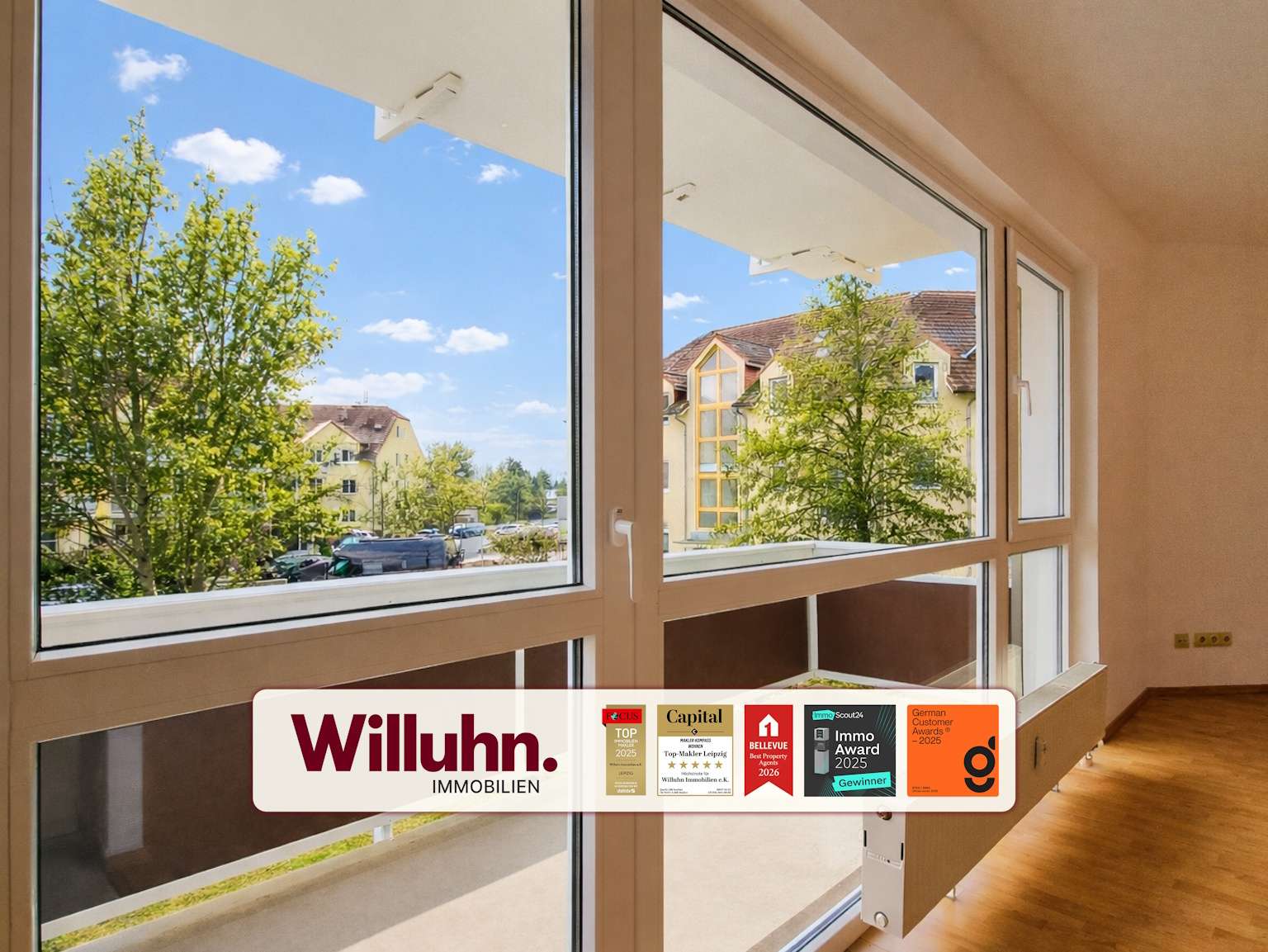 Bright living space with large glass windows showing a tree-filled street and yellow buildings outside; Willuhn Immobilien branding banner with awards along the bottom
