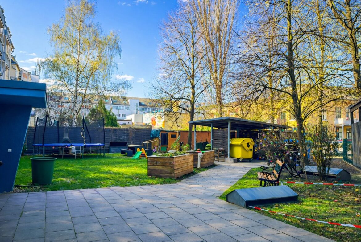 Urban backyard play area with a trampoline, slide, raised wooden planters, and a yellow recycling bin under a shelter near trees and apartments in the background.
