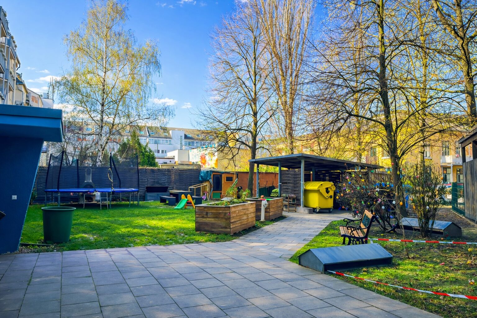 Urban backyard play area with a trampoline, slide, raised wooden planters, and a yellow recycling bin under a shelter near trees and apartments in the background.