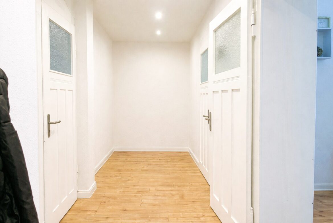 Bright white hallway with two white paneled doors on either side, frosted glass panels, and light wood flooring.”],