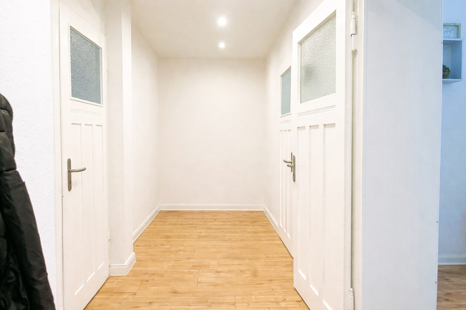 Bright white hallway with two white paneled doors on either side, frosted glass panels, and light wood flooring.”],