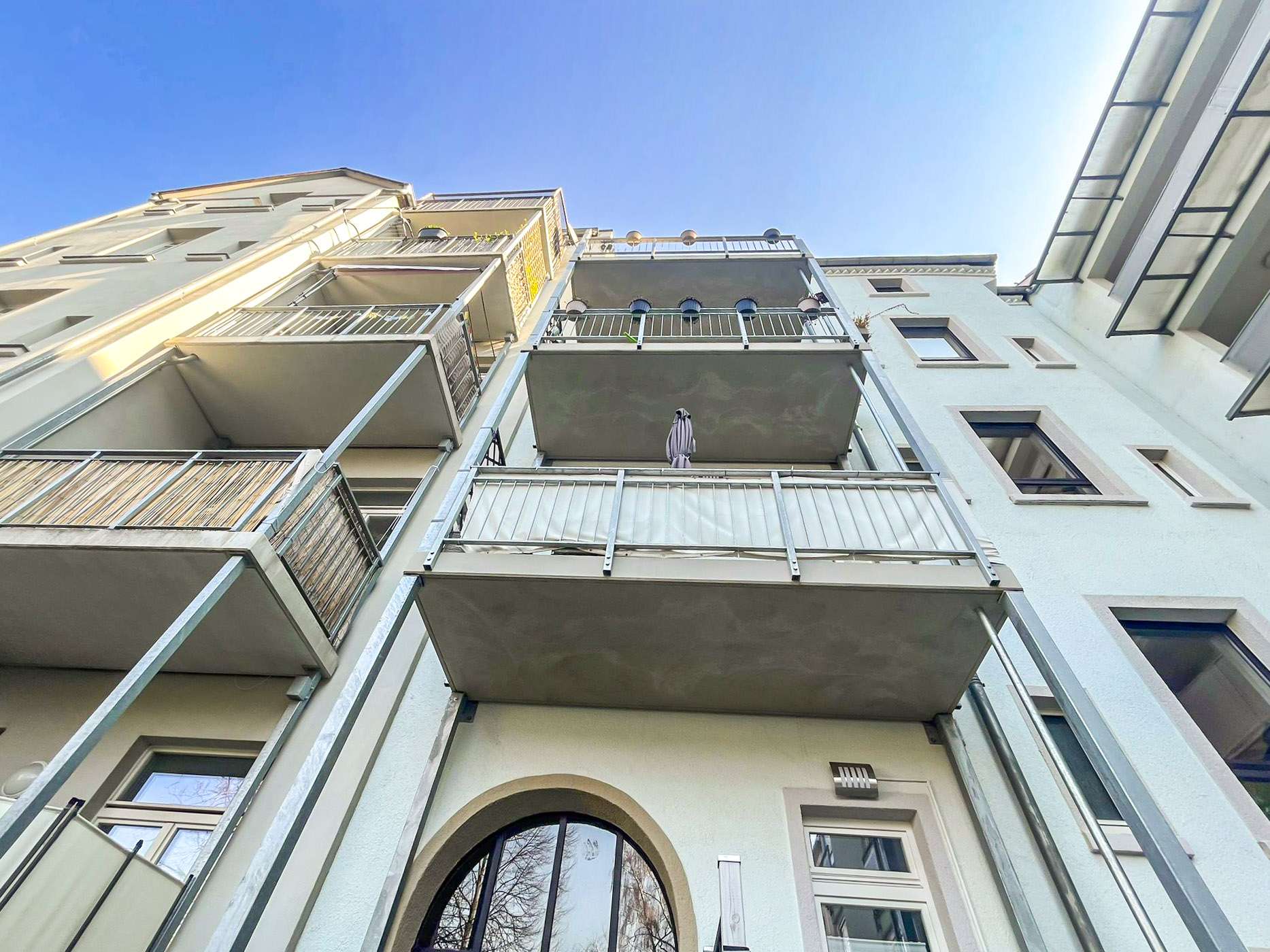 Low-angle view of a pale blue apartment building with balconies and glass railings against a clear blue sky, multiple floors visible and architectural details. (informational)