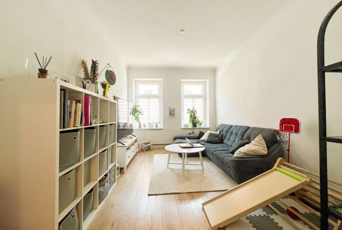 Cozy living room with a long gray sofa, white shelving unit, and large windows with plants on the sill.